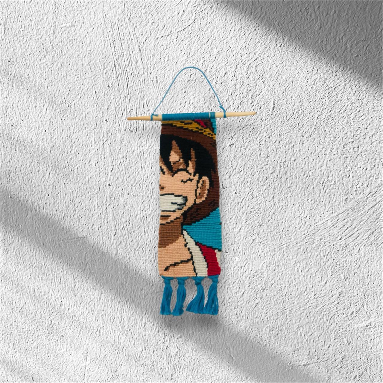 Handmade macramé wall hanging inspired by Luffy from One Piece, showing his wide smile, straw hat, and vibrant colors, with decorative blue tassels.