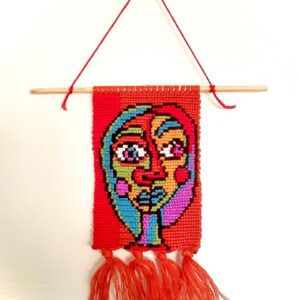 Handwoven red macramé wall hanging featuring an abstract multicolored face design with tassels, named Sti Zahra, hung on a wooden dowel.