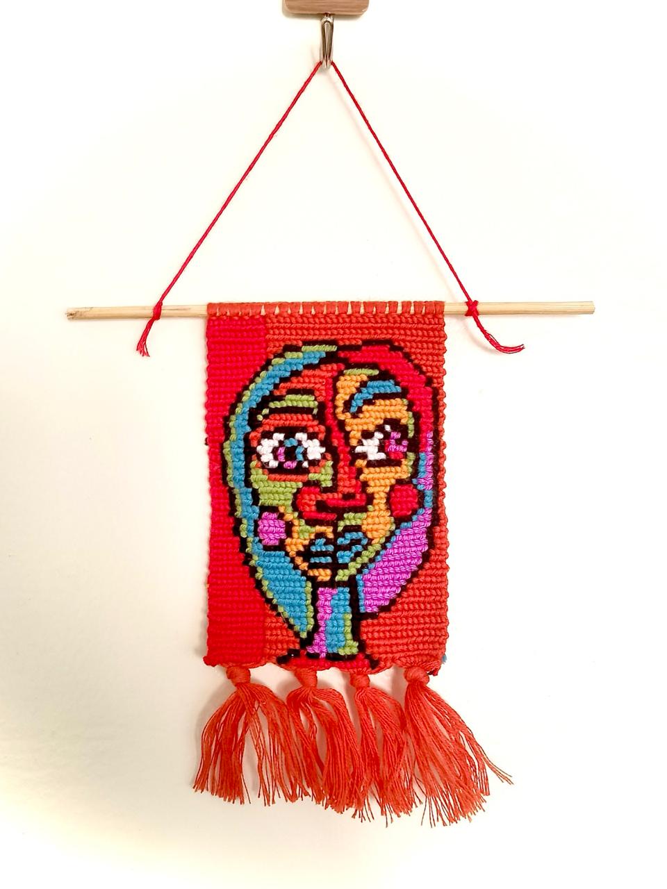 Handwoven red macramé wall hanging featuring an abstract multicolored face design with tassels, named Sti Zahra, hung on a wooden dowel.