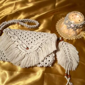 Handmade macramé shoulder bag with fringe flap, wooden bead accents, and matching woven strap displayed on golden fabric.